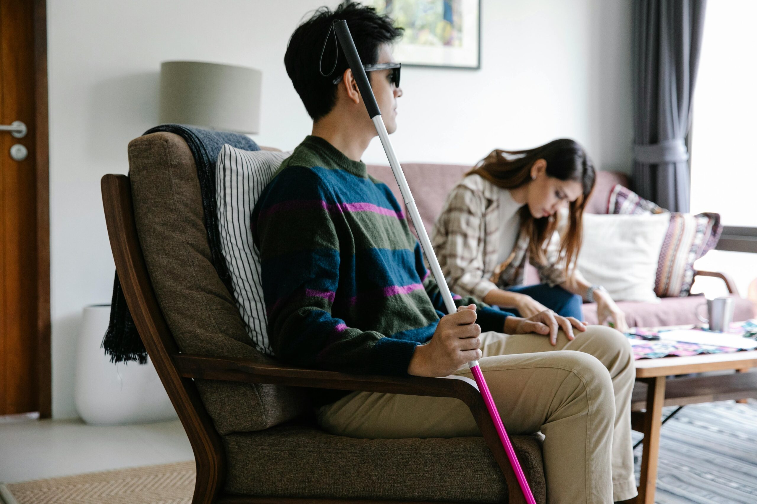 A blind man holding a cane sits in a cozy living room with a woman sitting nearby.